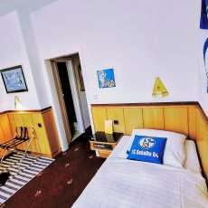 Ein einfaches Schlafzimmer mit einem weißen Bett, ausgestattet mit einem blauen Kissen der Schalke 04. An der Wand hängen Bilder, und ein großer Schalke 04-Wimpel ist sichtbar. Im Raum gibt es eine Tür und einen Tisch mit Stühlen.
