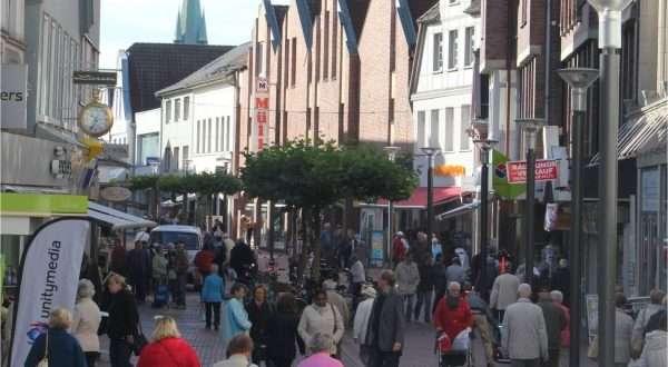 Eine belebte Fußgängerzone mit vielen Menschen, die entlang der Geschäfte und Restaurants spazieren. Bäume und Laternen sind in der Straße zu sehen, im Hintergrund ragt ein Kirchturm empor.