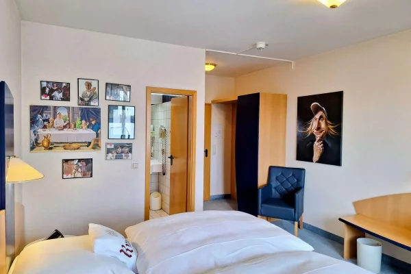 Ein Hotelzimmer mit einem Doppelbett, einem blauen Sessel, einem Tisch und Bildern an der Wand. Die Tür zum Badezimmer ist sichtbar, und ein Bild einer Person hängt neben einer Schranktür.